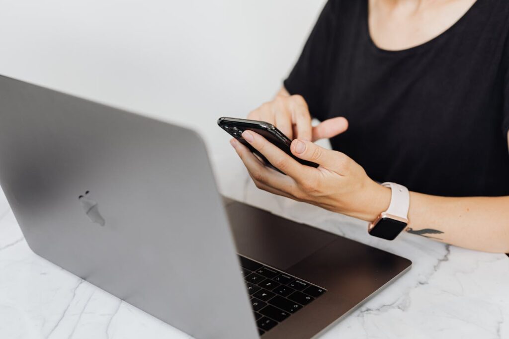 Close-up of woman using smartphone and laptop at desk, wearing a smartwatch.
